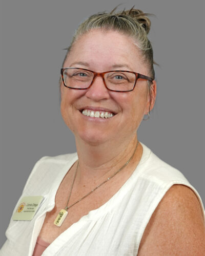 Connie Ortega has white and brown hair up in a bun, wears glasses and a white blouse and smiles for the camera.