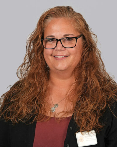 Shellanne Thompson has long red hair, is wearing square glasses and a black shirt, and smiles for the camera.