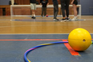 A yellow goal ball sits inside a blue, pink and yellow hula hoop on the floor of a gymnasium. People's feet and a goal ball net are visible in the background.